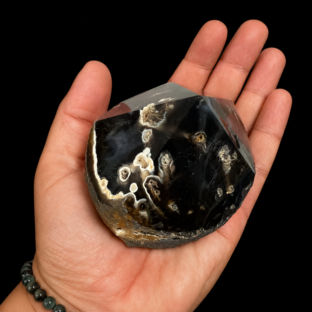 Hand-held front view of a unique black and cream semi-polished agate point with polished facets, natural cream inclusions and rough base, shown for size reference on a black background.