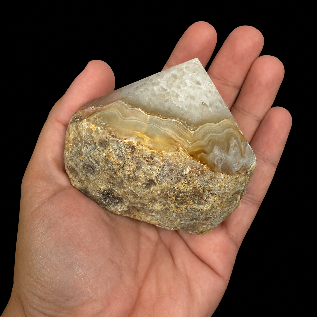 Hand-held angled front view of a unique cream honey semi-polished agate point with natural banding, rough base and polished apex, shown for size reference on a black background.
