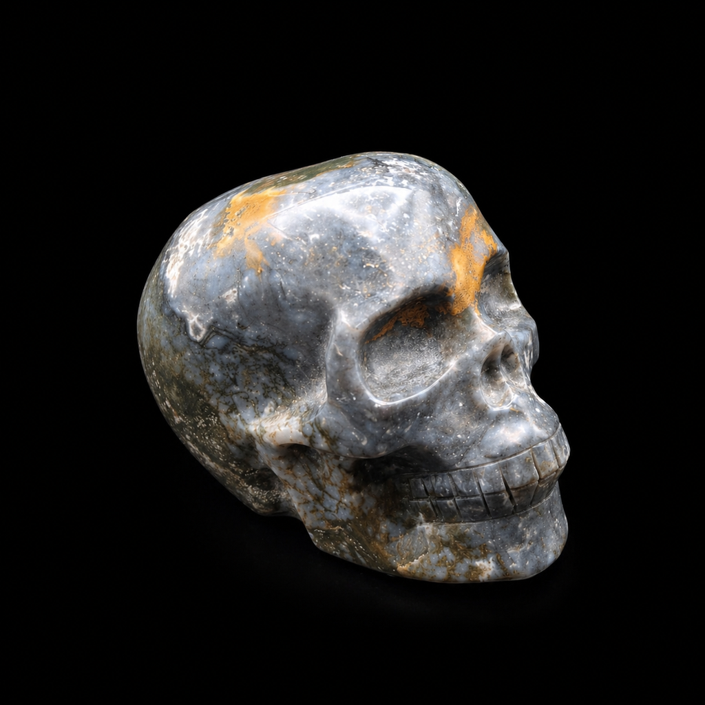Side view of a polished blue moss agate skull carving showing blue-grey agate patterns, golden mineral markings and carved facial detail