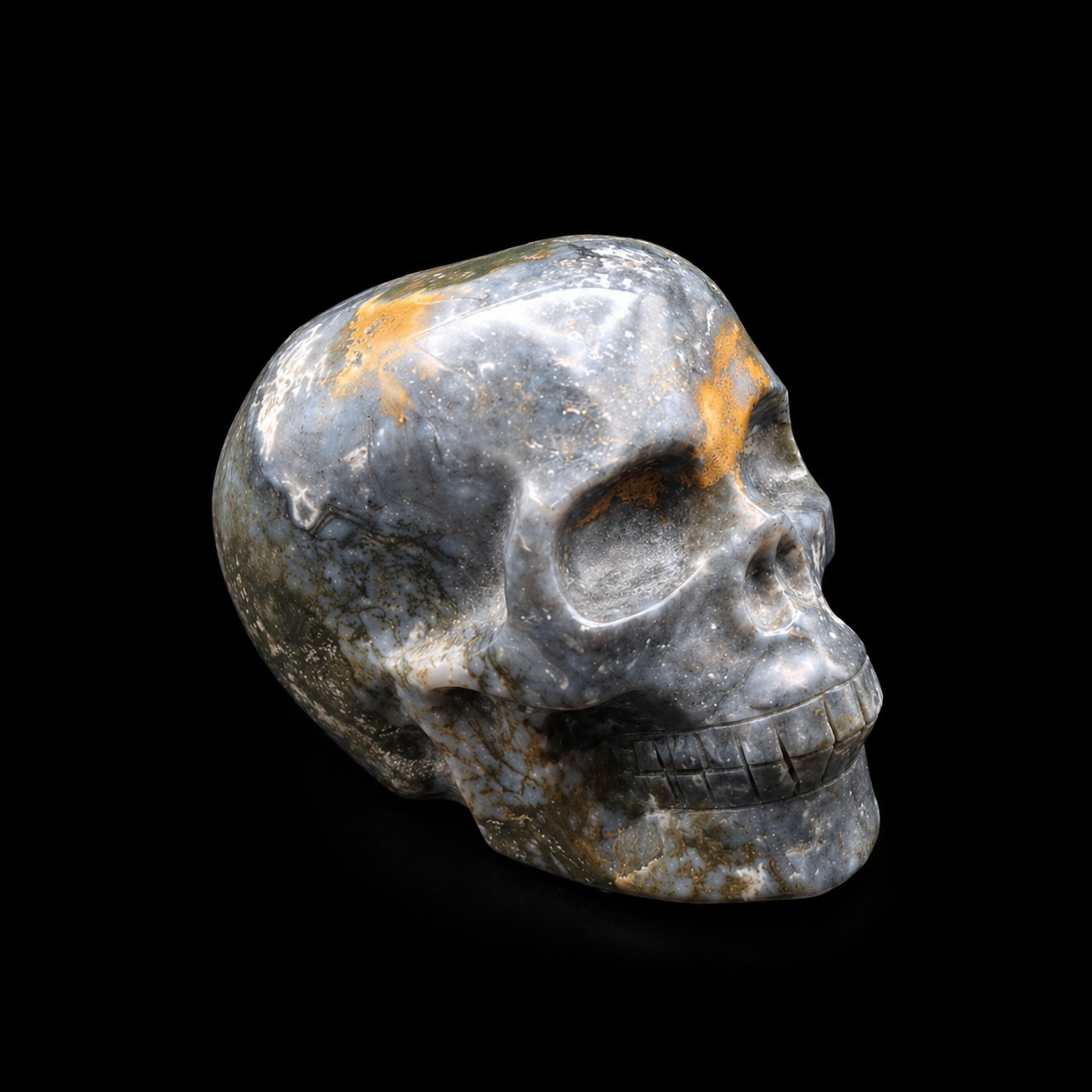 Side view of a polished blue moss agate skull carving showing blue-grey agate patterns, golden mineral markings and carved facial detail
