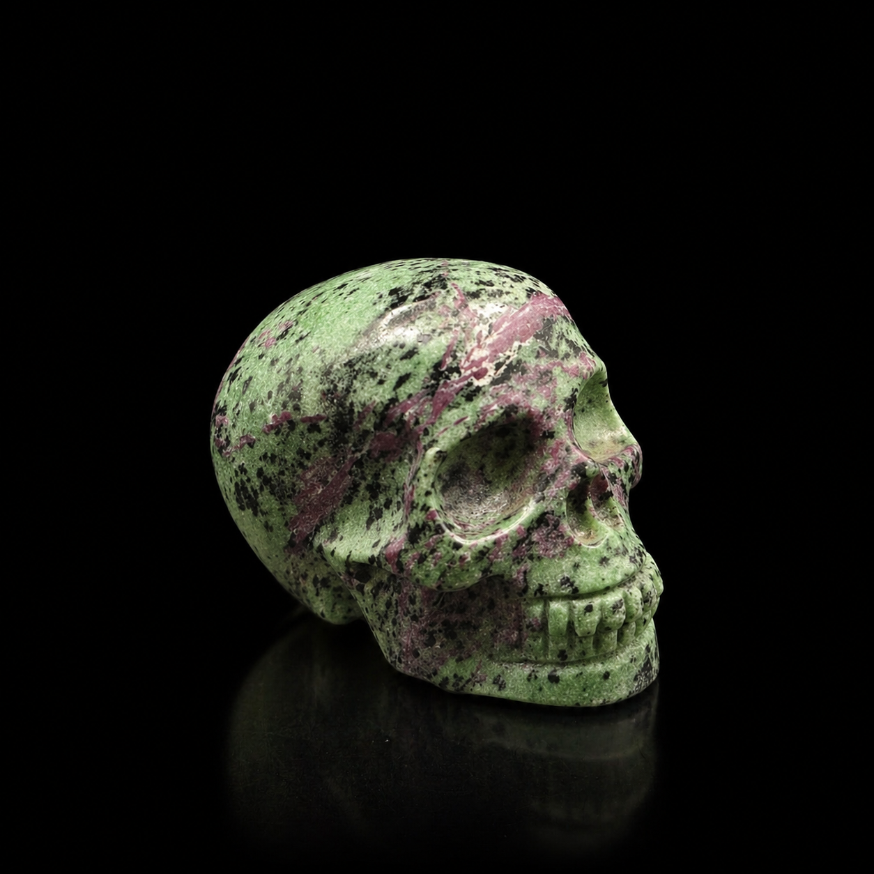 Ruby Zoisite skull angled side view, polished mineral carving with green zoisite, ruby-pink streaks and black inclusions