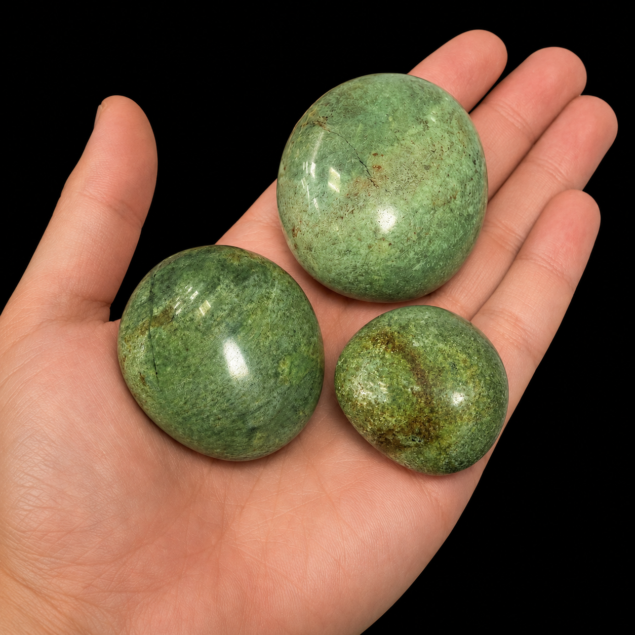 Three polished chrysoprase palm stones resting together on an open hand as a size reference, showing natural green tones, varied mottled patterns and glossy rounded surfaces against a black background