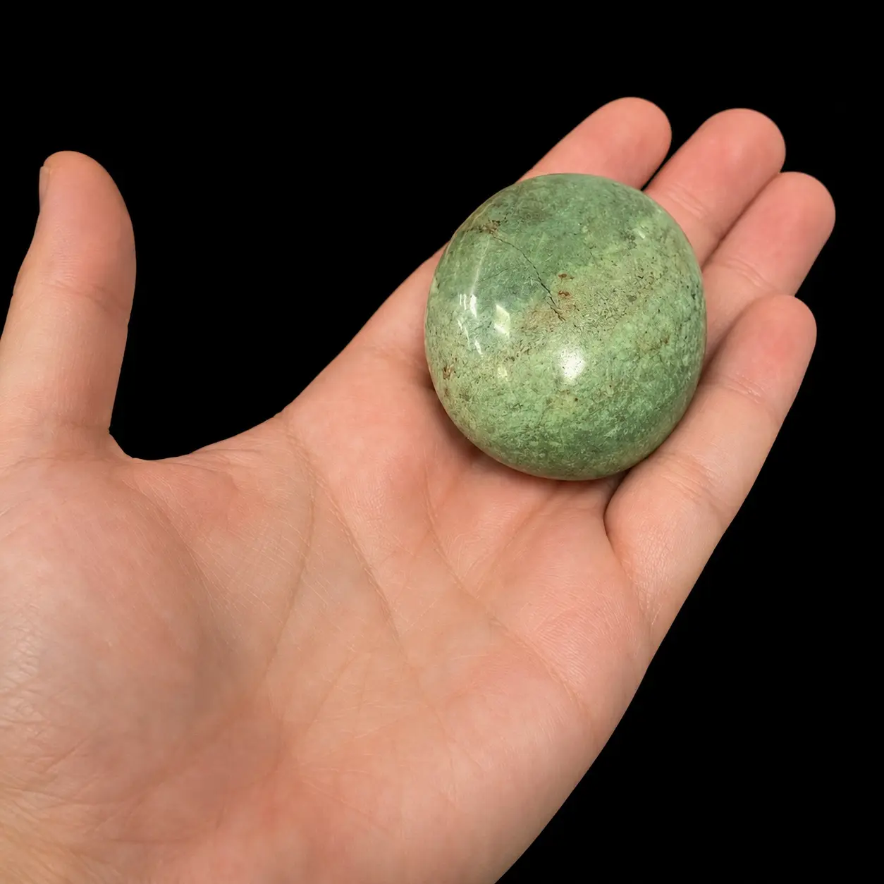 Polished chrysoprase palm stone resting on open hand as size reference, with soft green colour, natural speckled pattern and smooth rounded shape