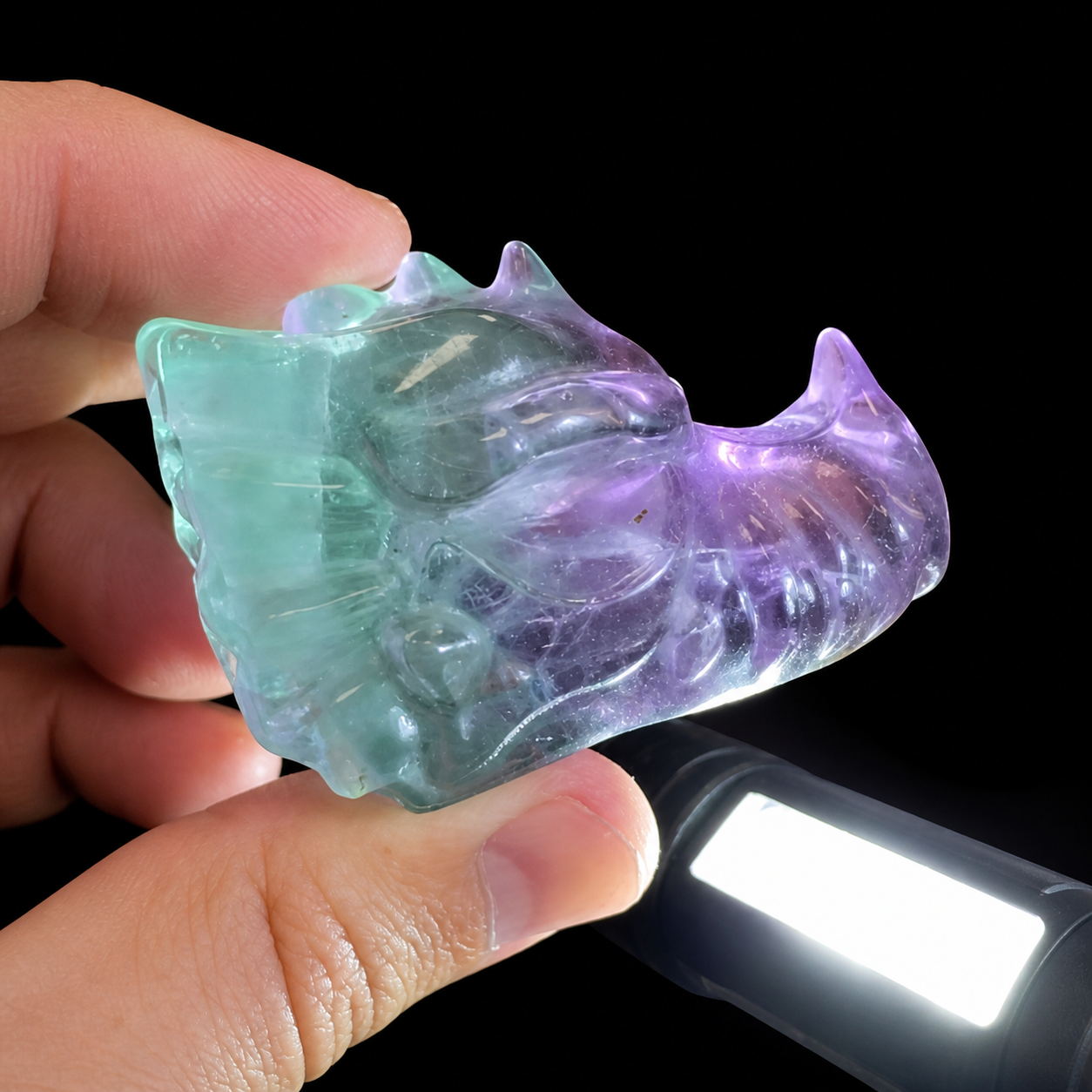 Fluorite dragon head carving held by hand with a flashlight showing translucent blue-green and purple natural colour zoning