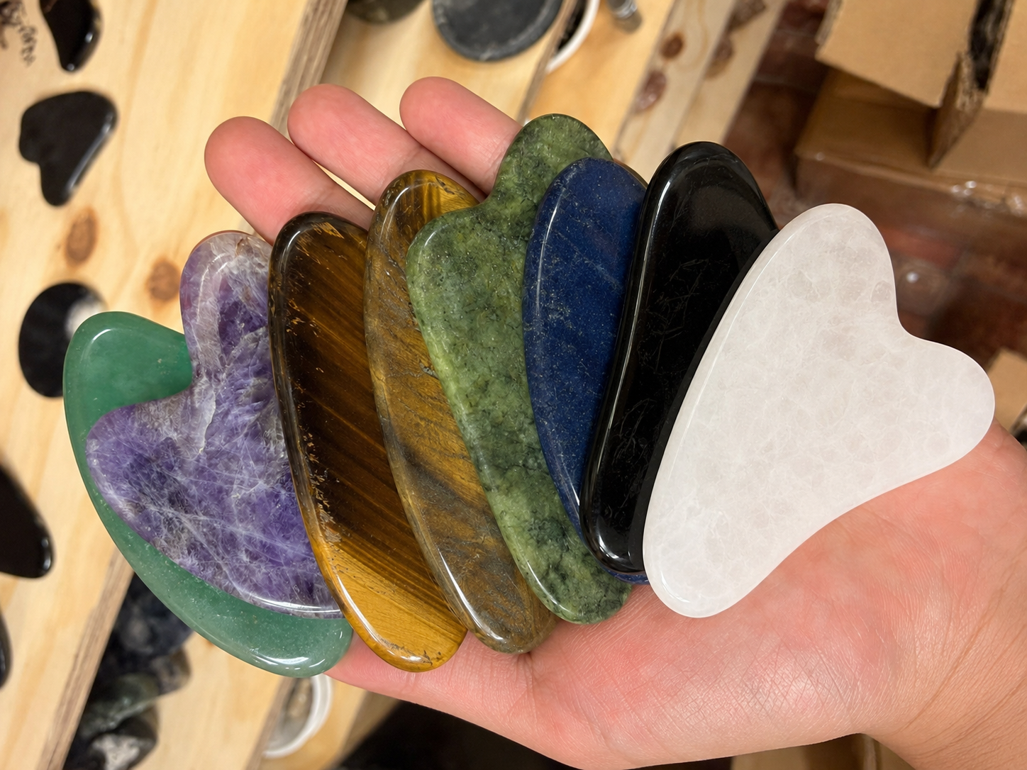 Hand holding assorted polished mineral Gua Sha tools, showing real size reference with white quartz, black obsidian, sodalite, green aventurine, tiger’s eye and amethyst