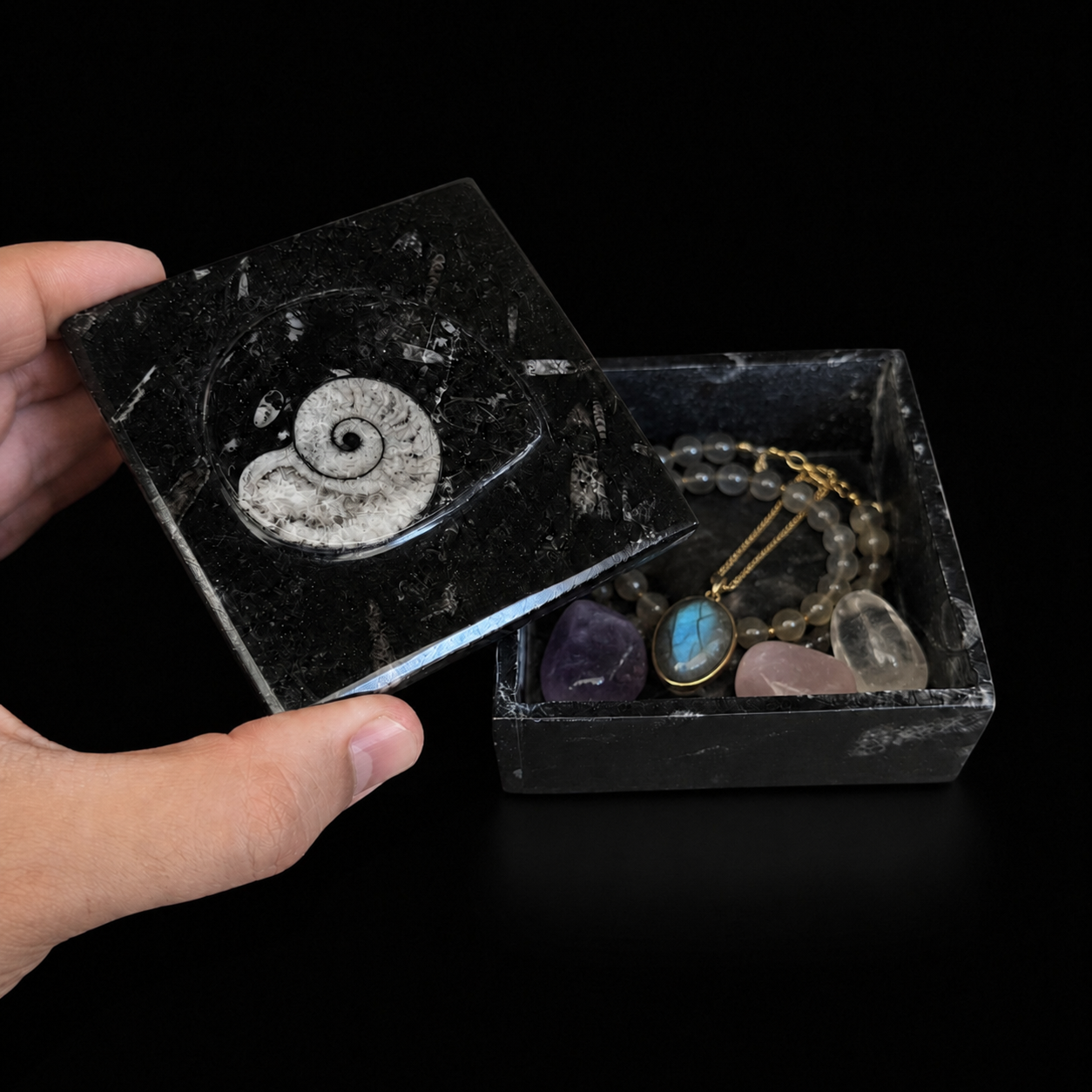 Open square fossil jasper jewellery box with lid held by hand, displaying a beaded bracelet, pendant necklace and polished mineral stones inside