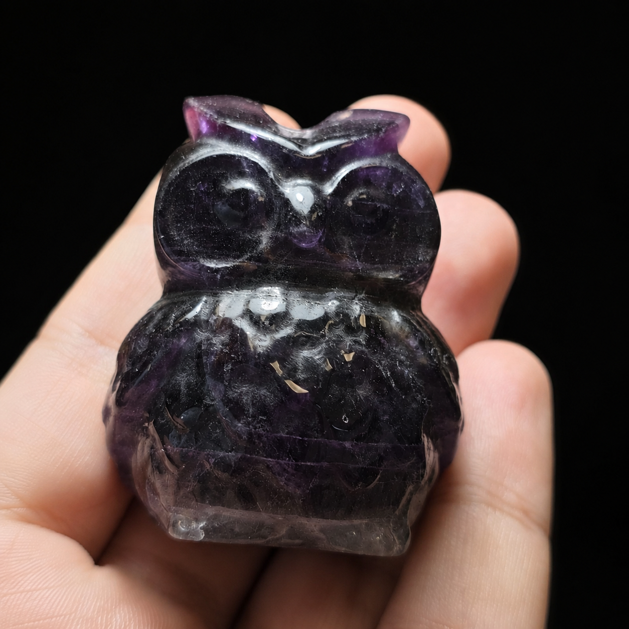 Front view of a small polished purple fluorite owl carving held in hand for size reference, showing dark violet tones, natural banding and carved details