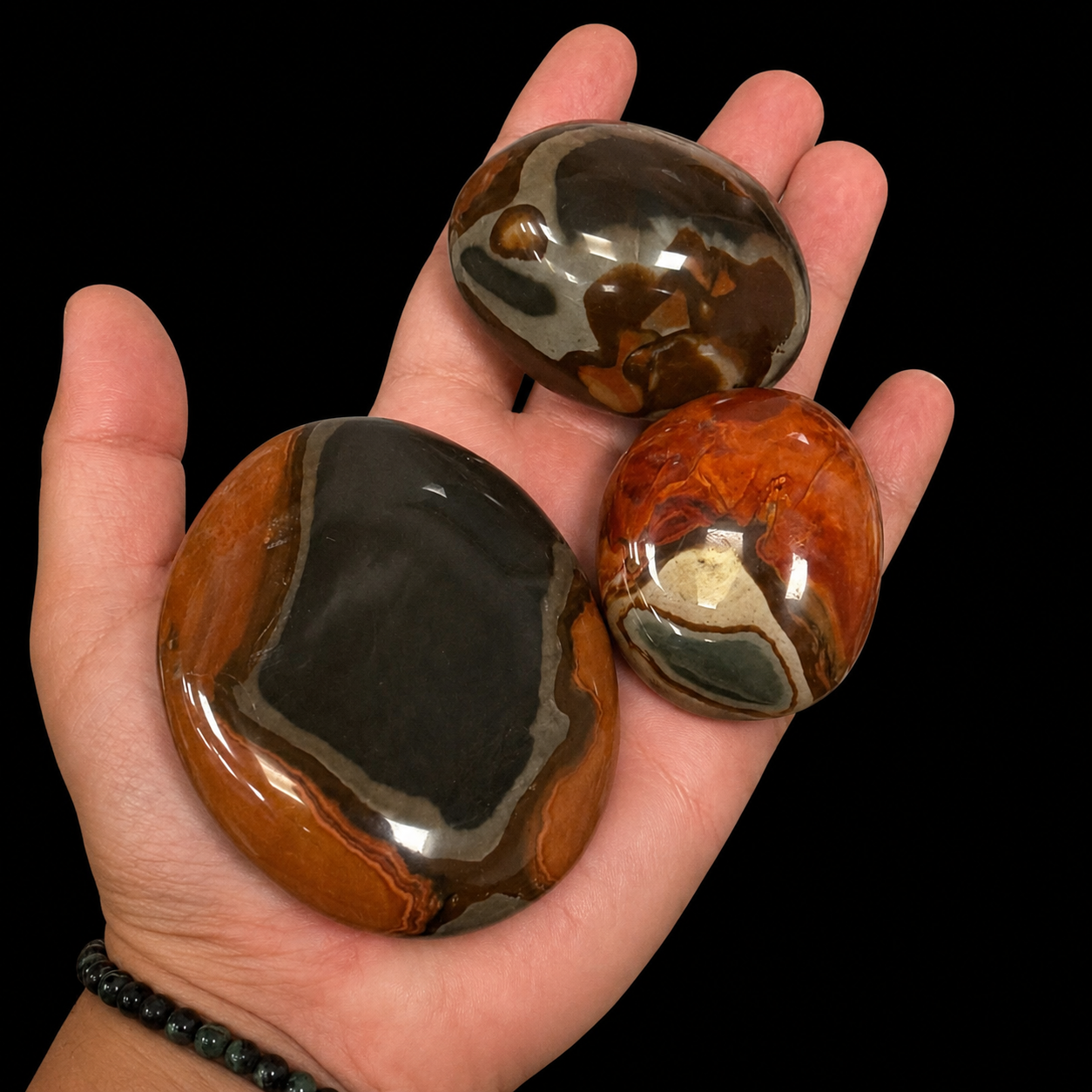 Polychrome jasper palm stones held in hand with earthy orange, grey and brown scenic patterns on a black background.