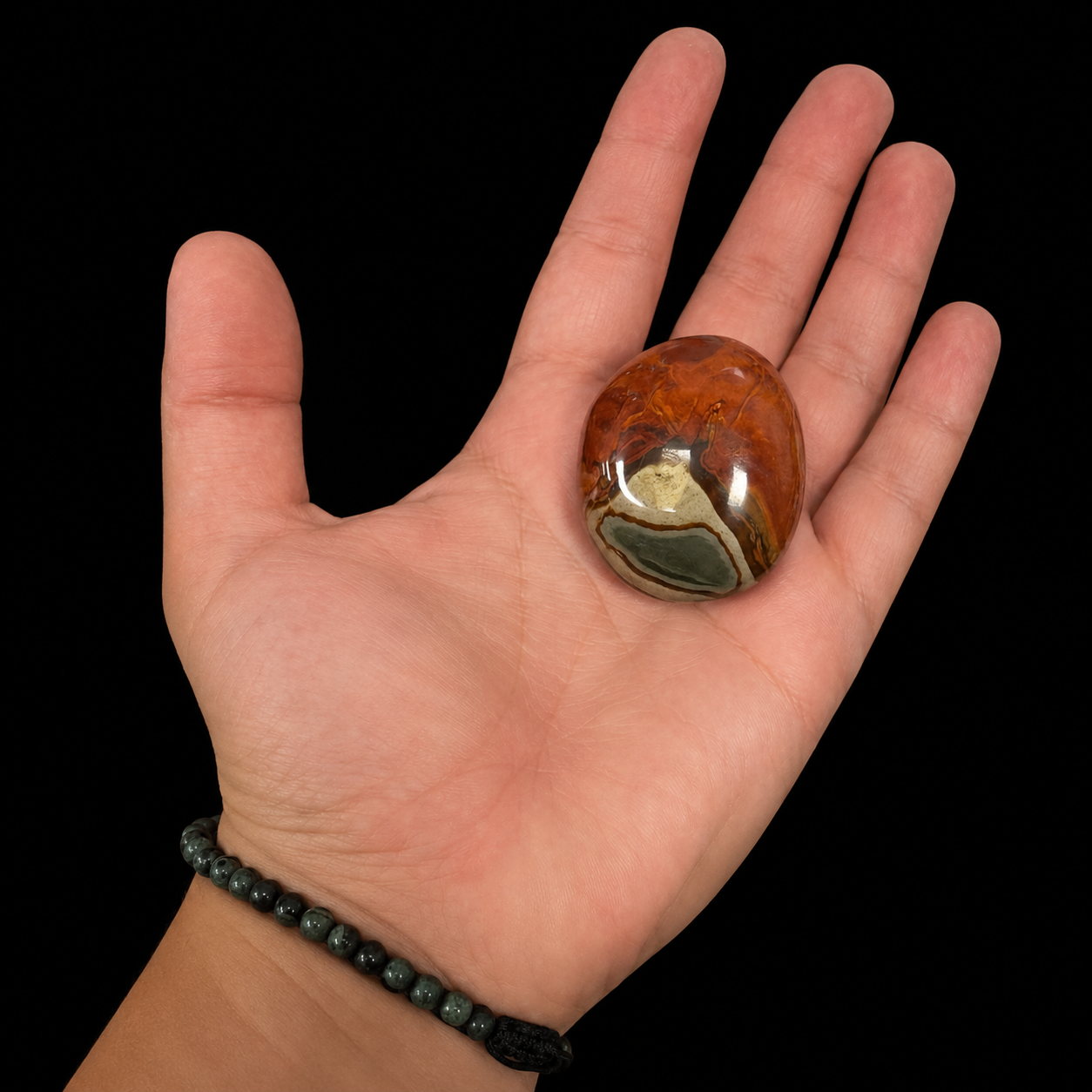 Small polychrome jasper palm stone held in hand, displaying vivid orange, cream and grey scenic patterns on a black background.
