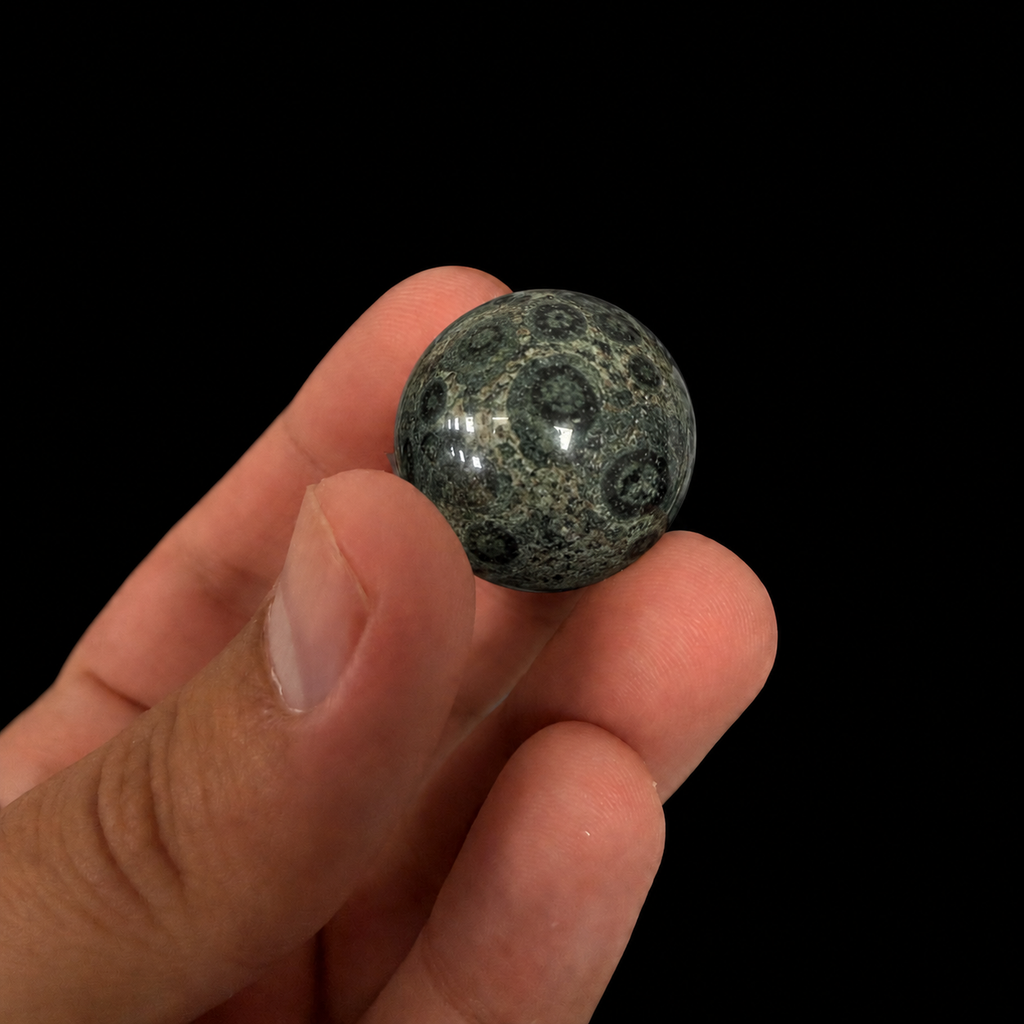 Single polished Kambaba mineral sphere held between the fingers, showing its rounded shape and natural circular patterning against a dark background.