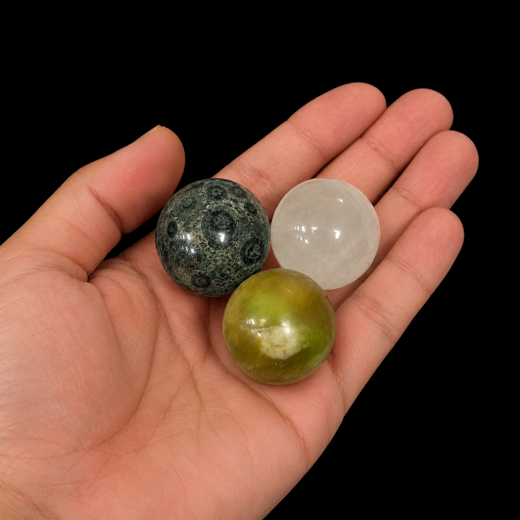 Three small polished mineral spheres in different varieties displayed in the palm, including Kambaba, translucent white and green, against a dark background.