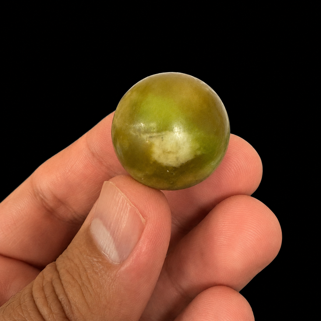 Single polished serpentinel sphere held between the fingers, showing its rounded shape and natural surface variation against a dark background.