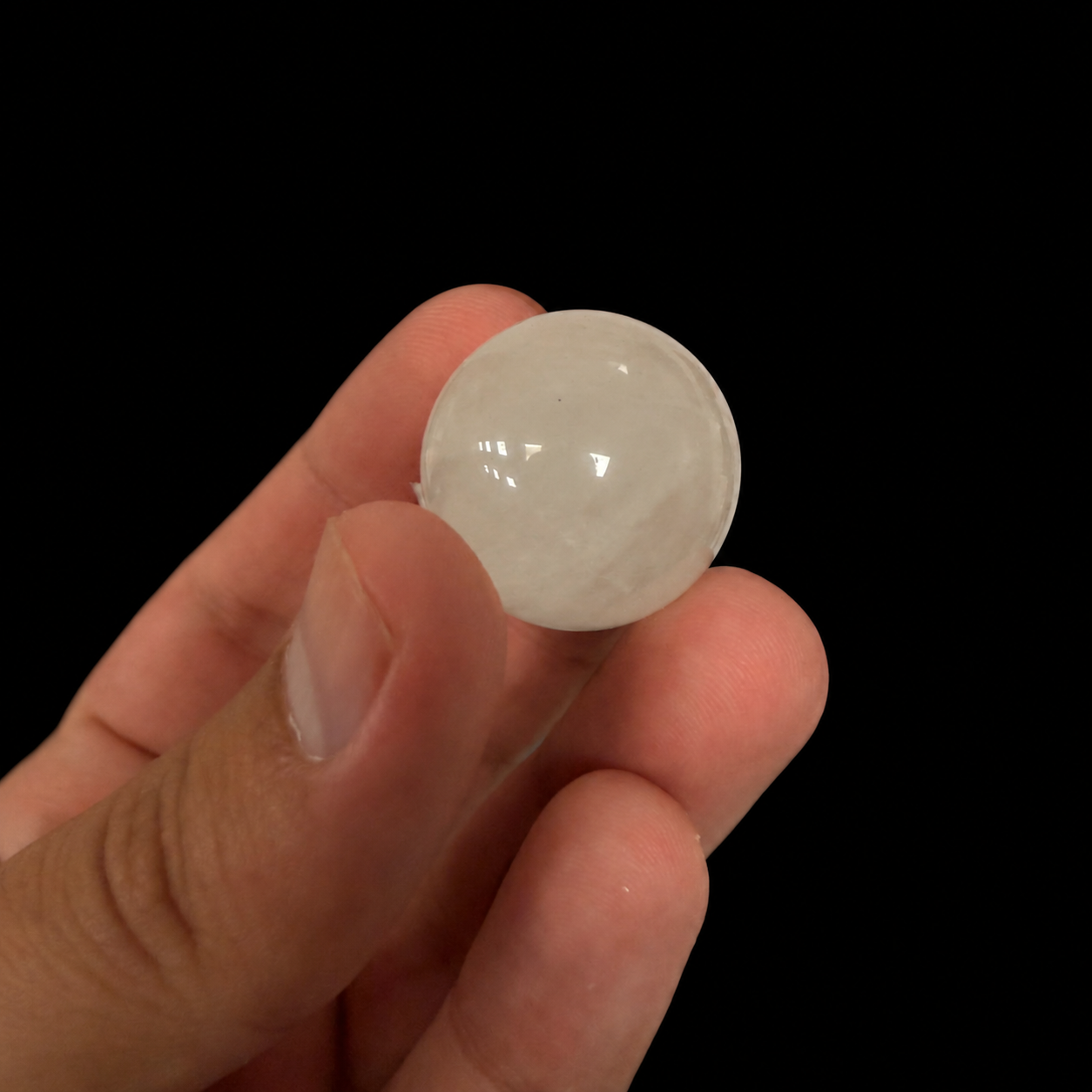 Single polished translucent white mineral sphere held between the fingers, showing its rounded shape and smooth glossy surface against a dark background.